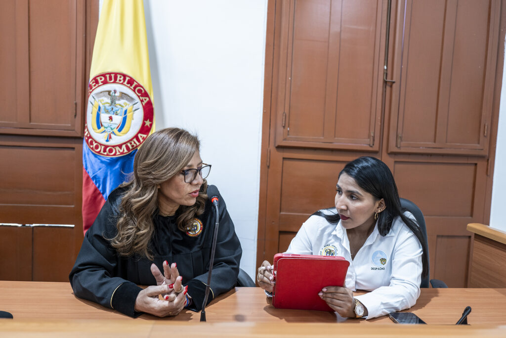 A woman in judges robes sits at a desk with a woman holding a tablet, with the Colombian flag in the background